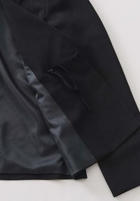 Zwarte blazer met een gladde satijnen voering. Bevat een gestructureerd ontwerp, textuur op de buitenstof en een verstelbare strik aan de binnenkant.