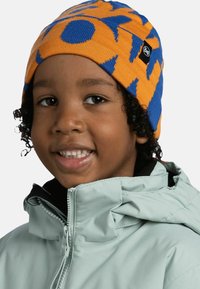 Knit beanie in orange with blue abstract patterns. Child's face visible, wearing a light green jacket with a high collar and zip.