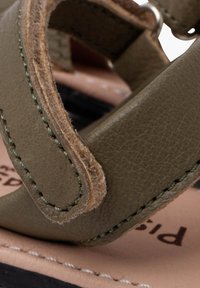 Tira de sanda verde de cuero con detalle de costura, que presenta una textura suave y una forma curva. Incluye un cierre metálico circular y una plantilla de cuero marrón claro.