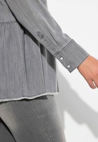 Camicia di chambray grigia con orlo sfrangiato, polsini con bottoni e texture morbida; abbinata a leggings in denim grigio, con attenzione ai dettagli delle cuciture.