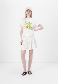 Fehér, crop fazonú póló színes citrom grafikával, fehér hímzett mini szoknyával és fekete flip-flop papuccsal párosítva. Csipke mintás kalap.