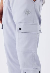Pantaloni cargo grigi chiari con tasche a pattina fissate da bottoni neri e un sottile logo ricamato sulla coscia.