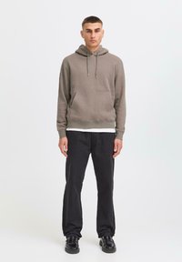 A young man stands in a light background, wearing a brown hoodie, black pants, and black shoes, with hands resting at his sides.