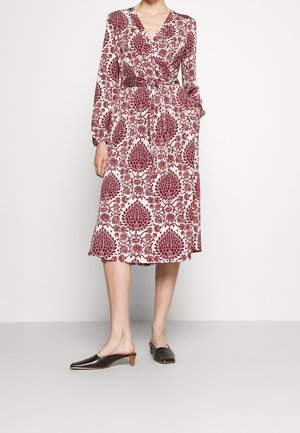 Kobieta ubrana w midi kopertową sukienkę z długim rękawem w czerwono-biały kwiatowy wzór oraz czarne mule na niskim obcasie, stoi z jedną ręką w kieszeni.