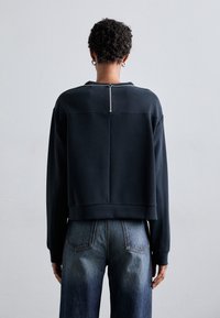 Sudadera negra corta con cuello alto, detalle de cremallera en la parte posterior y puños acanalados. Combinada con jeans de denim azul de cintura alta.