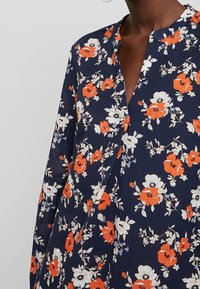 Blusa navy floreale con fiori rossi e bianchi, maniche lunghe e colletto a V. Tessuto leggero con una vestibilità ampia e stampa vivace.
