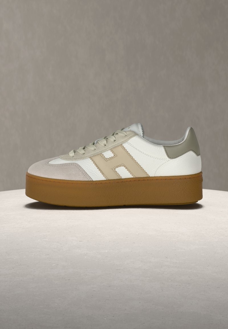 Baskets compensées blanches et beiges avec une semelle épaisse marron, un design à lacets, et un logo "H" beige cousu sur le côté, présentées sur une surface neutre.