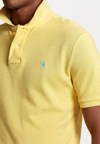 Yellow polo shirt made of textured cotton, featuring a two-button placket, short sleeves, and an embroidered blue logo on the chest.