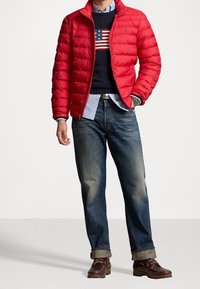 Červená péřová bunda, zapínání na zip, prošívaný design. Námořnický svetr s vzorem americké vlajky, modrá košile na knoflíky a vybledlé džíny. Hnědé boty.