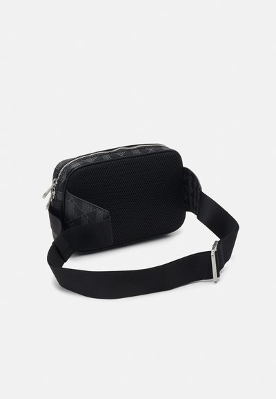 Sac rectangulaire noir avec une surface en maille texturée, des fermetures éclair argentées et une sangle noire réglable. Présente un motif géométrique subtil sur un côté.