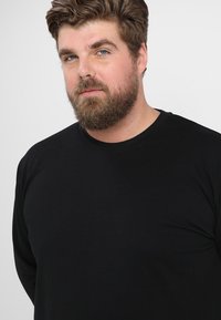 Must tee-särk, millel on pikad varrukad ja mis on valmistatud pehmest puuvillast. Särgil on ümmargune kaelus ja lõdva istuvusega, minimaalsete õmbluste detailidega.