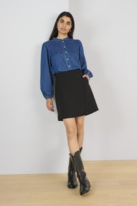 Chemise en denim à manches bouffantes, associée à une jupe portefeuille noire et des bottes noires montantes sur un sol en bois clair.