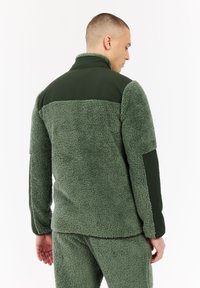 Hombre vistiendo una chaqueta de forro polar sherpa verde oliva y pantalones a juego, de espaldas contra un fondo blanco liso.