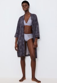 Donna che sta scalza in lingerie di pizzo lilla e in un accappatoio di colore viola scuro con motivo floreale, su uno sfondo bianco semplice.
