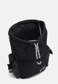 Svart tygryggsäck med dragsnörning, grå rutiga detaljer på sidorna och reflekterande beslag. Inredningen är klädd i ljusgrått.