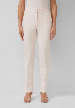 Kobieta nosząca jasnoróżowe, wysokie w talii, dopasowane spodnie z przodu z kantem, zestawione z białymi, paskowanymi sandałami na wysokim obcasie na jednolitym tle.