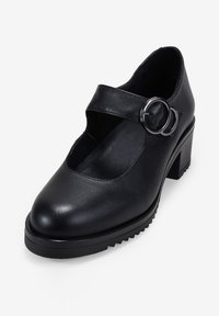 Zapato Mary Jane de cuero negro con una hebilla plateada redonda, tacón grueso y suela texturizada sobre un fondo blanco.