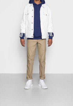 Man staat met een witte denim jack, een marineblauwe hoodie met gestreepte manchetten, beige broek, witte sokken en witte sneakers.