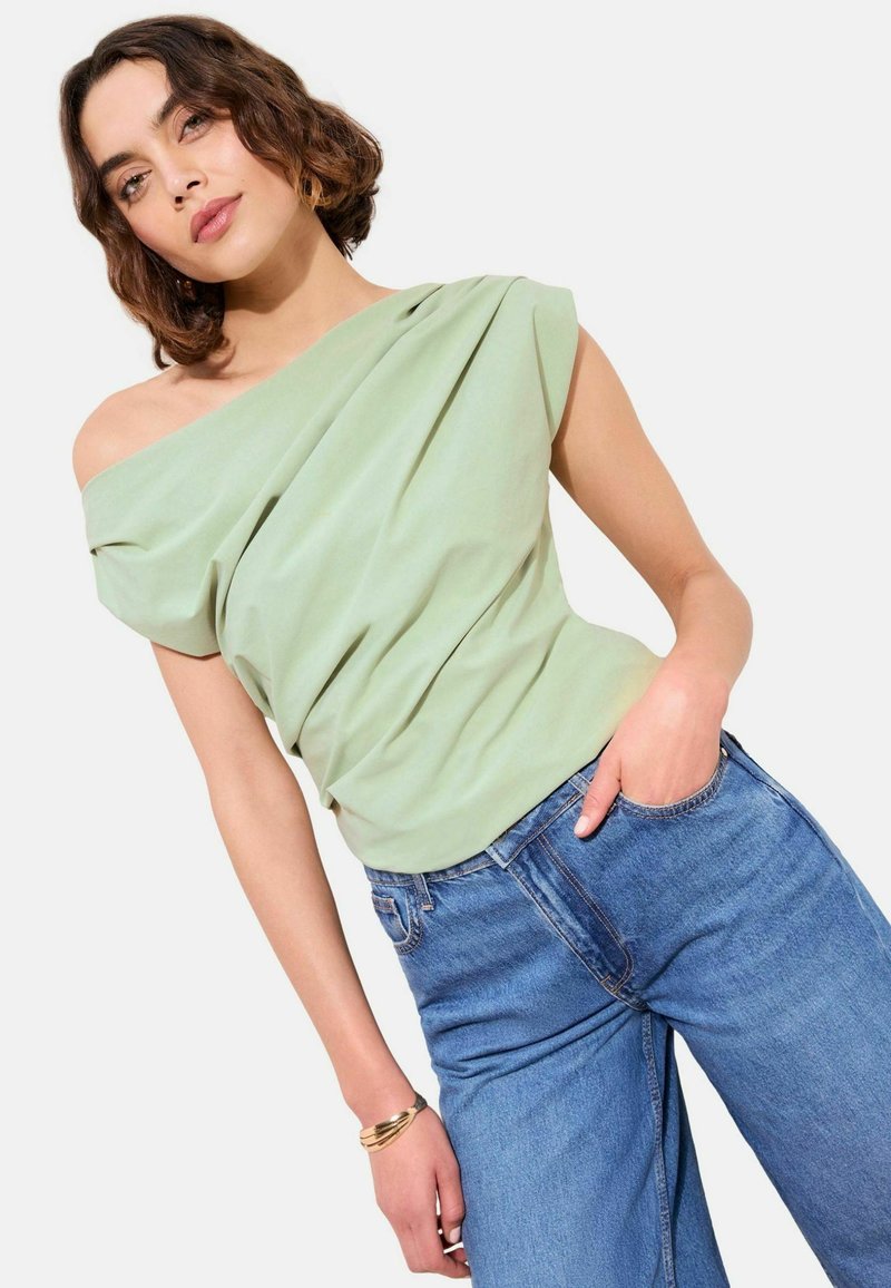 Vrouw met kort golvend haar, gekleed in een lichtgroene een-schouder gedrapeerde top en een blauwe spijkerbroek, hand in de zak, poseert tegen een effen achtergrond.