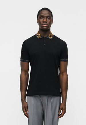 Férfi fekete rövid ujjú galléros pólóban arany szegélyű gallérral és ujjvégekkel, világosszürke redős nadrággal párosítva.