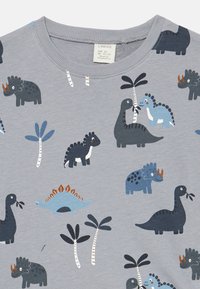 T-shirt grigia in cotone con stampe colorate di dinosauri e palme in blu, blu scuro e nero. Collo a giro, texture morbida, design giocoso.