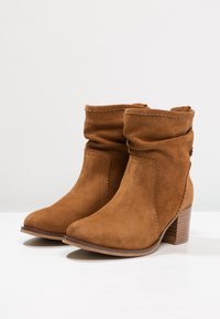 Tan mockasins med en slapp design, rund tå och klack i stapel, med dekorativ sömnad längs kanterna.