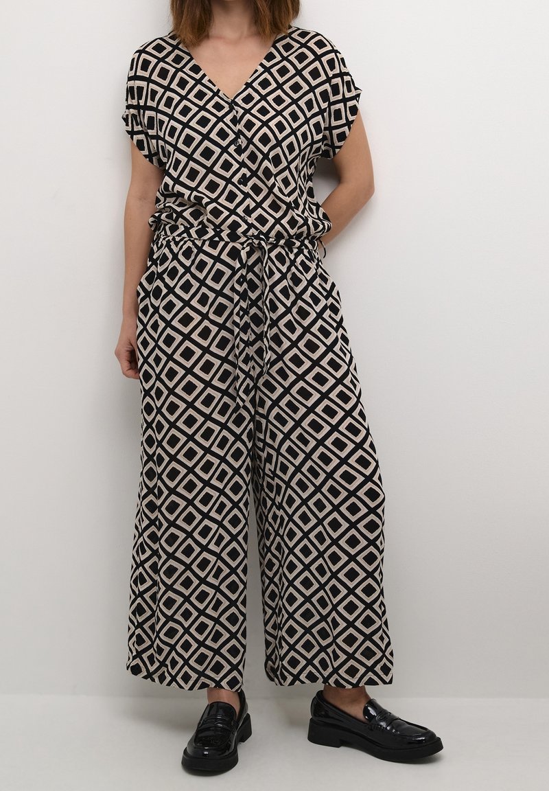 Femme portant un haut et un pantalon large à motif géométrique noir et beige assortis, ainsi que des mocassins noirs brillants, devant un mur blanc uni.