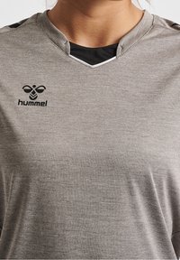 Szara koszulka sportowa z dekoltem w serek, czarnymi akcentami oraz małym czarnym logo Hummel na piersi. Wykonana z oddychającego materiału o teksturowanym wykończeniu.