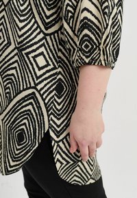 Schwarz-creamfarbige Bluse mit geometrischen Mustern, gerafften Ärmeln und lockerer Passform; getragen über schwarzer Hose, die Textur und Details zur Schau stellt.