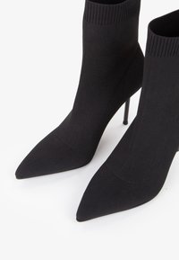 Czarne dzianinowe botki z wydłużonym noskiem i cienkimi wysokimi obcasami na białym tle.