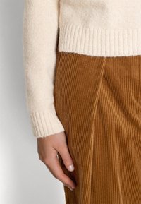 Main d'une femme posée contre un pantalon en velours côtelé marron taille haute, portant une manche de pull en maille côtelée de couleur crème.