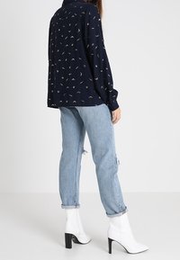 Donna che indossa una blusa a fantasia blu navy, jeans azzurri arrotolati e stivaletti bianchi con tacco alto, in piedi contro uno sfondo semplice.