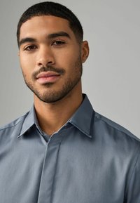 Camisa de manga corta de botones en un azul claro, confeccionada con una tela suave; presenta un cuello estructurado y un sutil pliegue frontal.