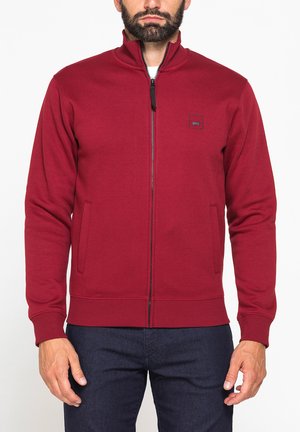 Uomo che indossa una giacca bordeaux con zip e collo alto e pantaloni scuri, in piedi contro uno sfondo bianco uniforme.