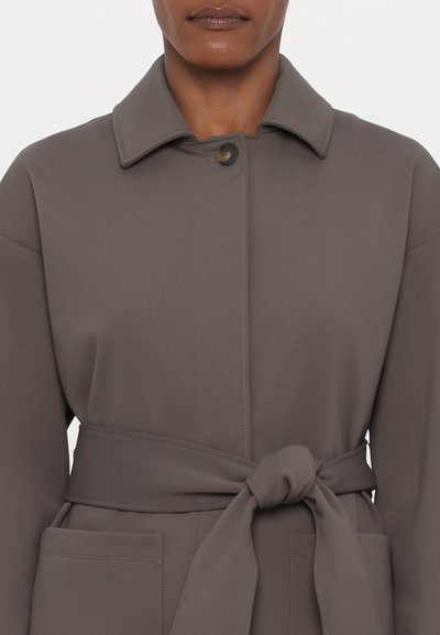 Grau mit Gürtel versehenes Mantel mit kragenlosem Ausschnitt, ausgestattet mit Fronttaschen und Steppnähten. Hergestellt aus glattem, strukturiertem Stoff.