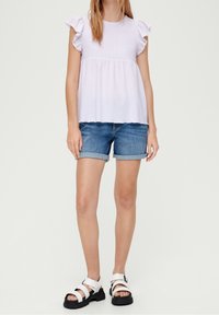 Hellblaue Bluse mit Rüschensäumen und gerafftem Oberteil. Kombiniert mit Jeans-Shorts mit umgeschlagenen Säumen. Die Schuhe sind weiße Plattformsandalen mit Riemen.