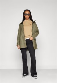 Olivengrønn vanntett jakke med hette, beige genser med lapp, svarte flagrende jeans og sorte joggesko. Solbriller fullfører antrekket.