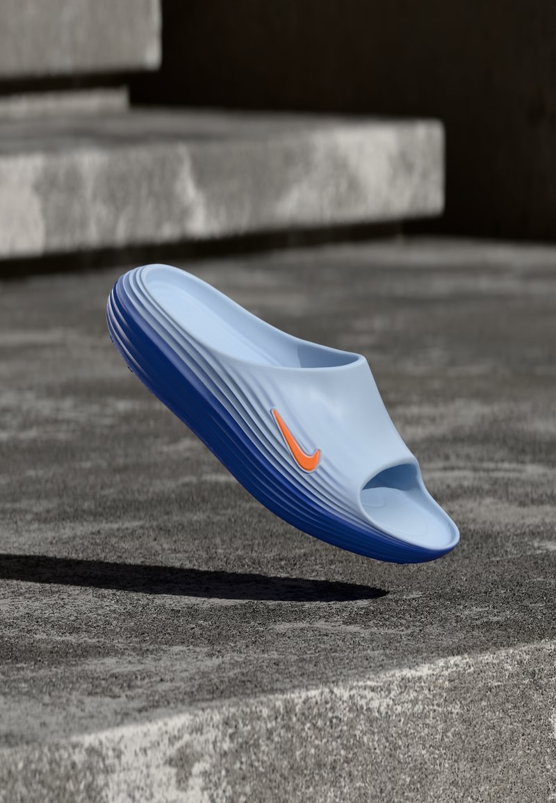 Sandale à glissière bleu clair et marine avec un logo Nike orange, en plein air au-dessus d'une surface en béton avec des marches en arrière-plan.