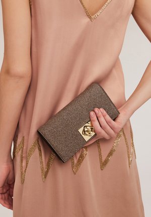 Braune Glitzer-Clutch mit goldenen Beschlägen und einem floralen Akzent, gehalten gegen ein pinkes Kleid mit goldenen Zickzackmustern.