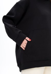 Felpa nera con cappuccio, dotata di tasca a marsupio, polsini a coste e una texture morbida, mostrata parzialmente con una mano infilata nella tasca.