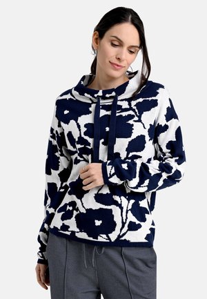Navy and white floral-patterned pullover hoodie with a wide neckline, drawstrings, and soft texture. Paired with gray pants.