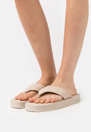 Des tongs beiges avec une semelle épaisse et de larges brides croisées, fabriquées en matériau lisse et flexible. Le design présente une forme de orteil arrondi.