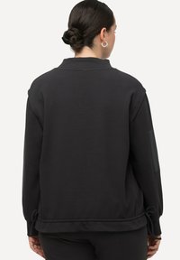 Felpa nera con finitura testurizzata, vestibilità oversize e cordoncino regolabile in vita. Presenta un'applicazione a patch sulla manica.