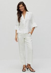 Witte linnen blouse met een V-hals en 3/4 mouwen, gecombineerd met bijpassende ruime linnen broeken. Model draagt strappy zwarte sandalen.