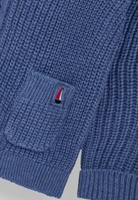 Pulover tricotat de culoare albastru închis, cu textură groasă. Dispune de o buzunar aplicat și un logo mic cu o barcă cu pânze roșie pe un fundal marin.