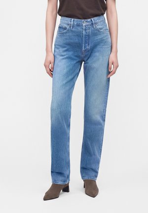 Person iført lyseblå højttaljede jeans med lige ben og brune ankelstøvler med spidse tæer, står mod en ensfarvet baggrund.