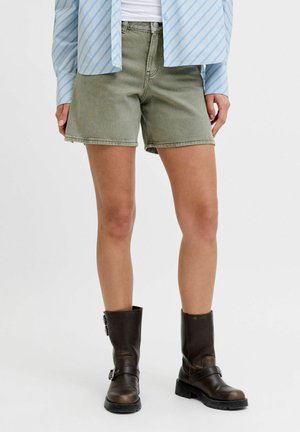 Személy olíva zöld farmer rövidnadrágot, világoskék csíkos gombos inget és sötétbarna térdig érő csizmát visel csatokkal.