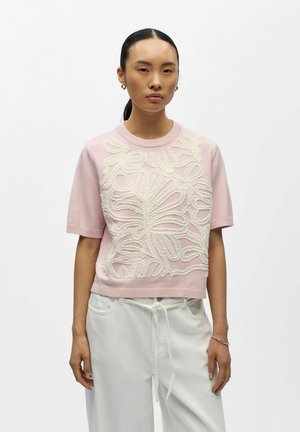 Donna con capelli neri lisci raccolti indossa un maglione rosa a maniche corte con ricami floreali bianchi e pantaloni bianchi larghi con coulisse.