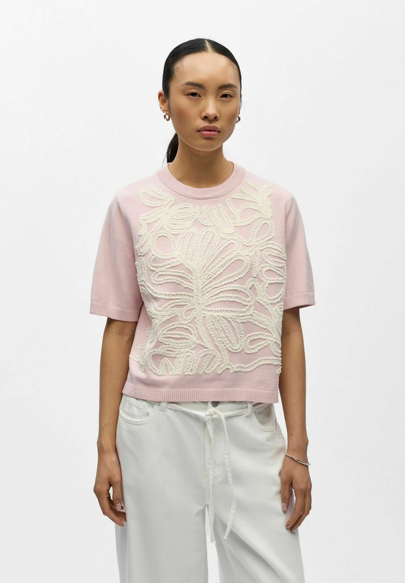 Femme aux cheveux noirs raides attachés derrière, portant un pull à manches courtes rose avec une broderie florale blanche et un pantalon blanc ample avec un cordon de serrage.