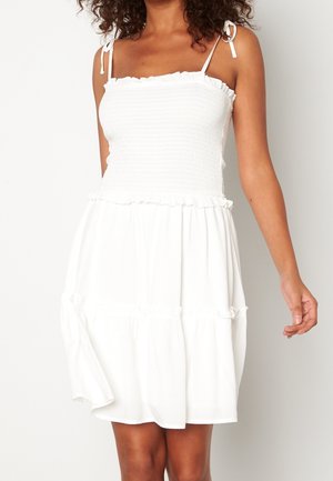Femme portant une robe blanche sans manches avec un corsage smocké et des bretelles à nouer, debout devant un fond clair uni.